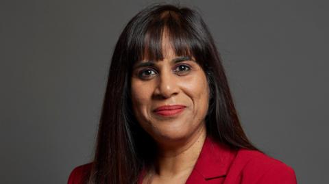 The MP Sureena Brackenridge is looking directly at the camera. The background is grey. She has dark long hair and is wearing a red jacket.