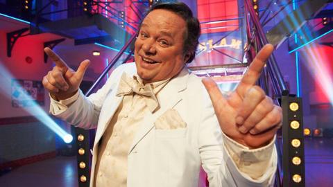 Ted Robbins in a cream tuxedo, waistcoat ad bow tie in a brightly lit set with spotlights. He has short dark hair and is smiling at the camera with his hands raised and index fingers pointing in the air