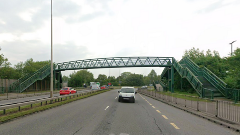 Spytty Road, a multi-lane road runs beneath a green metal pedestrian footbridge with staircases on both sides. Several cars and a white van travel along the road, and trees line both sides.