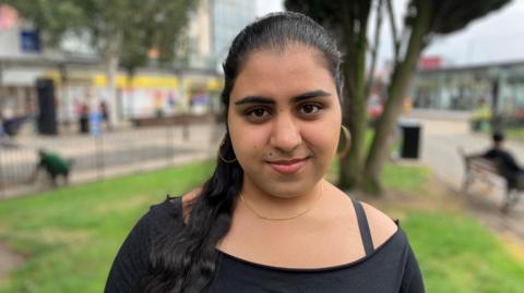 Safi smiles while standing in an outdoor area. She is wearing a black top and has long black hair tied in a ponytail. 
