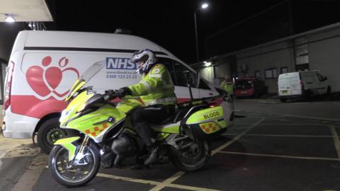 A motorcyclist in a high vis jacket on a high vis motorbike pulling in to park the bike next to an NHS blood van. His bike has "blood" clearly visible on the rear of the vehicle.