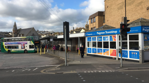 The image shows a bus station in Paignton, Devon. In the foreground, there is a pedestrian crossing with traffic lights and a road marking that says “LOOK LEFT.” To the right, there is a building with a bright blue sign reading “Paignton Travelshop,” and its windows are covered with posters advertising travel services and ticket information. In the centre and left of the image, there are buses parked, including a green and white double-decker bus with an advert on its side. Behind the buses, there is a covered walkway where people are standing. In the background, you can see rooftops of houses and a church with a tall spire, set against a cloudy sky with patches of blue. 