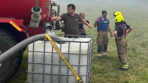 A man holds the controls of a large red tanker as it fills a plastic tank full of water. In the background, firefighters wearing high-viz helmets talk.