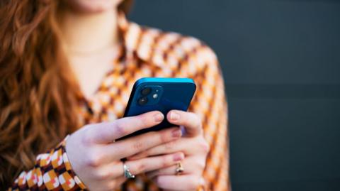 A woman holds a metallic blue coloured mobile phone in both her hands. 