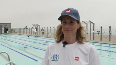 A woman wearing a white t-shirt and blue hat stood in front of a swimming pool. The pool is next to a rocky beach, with the sea in the background.