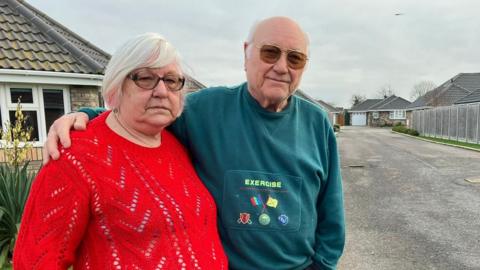 An older man and women stand side-by-side. The man has his arm around the woman's shoulder. The man is wearing a green jumper, and the woman is wearing a red jumper.  They stand on a road with bungalows. The sky is overcast and grey.