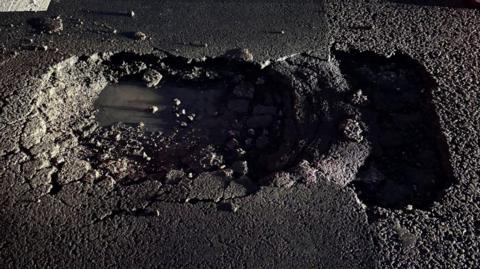 A close-up view of a damaged section of asphalt road surface. At the centre of the image, there is an irregularly shaped pothole with rough, crumbling edges where the top layer of the road has broken away. The inside of the pothole appears dark and damp. Loose stones and fragments of asphalt are visible around the edges of the hole. 