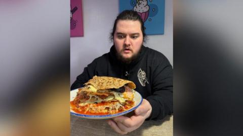 A man with long hair, a beard and wearing a black hoody, holds a plate with a massive oatcake on it full of food. He has a shocked expression on his face.