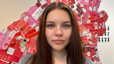 A young woman with long brown hair and a grey V-neck jumper stands in front of a section of the sculpture covered in flattened, red vape packets.