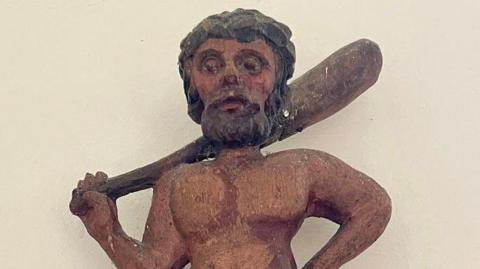 Close up of a wooden carving depicting a wild man. He has textured brown shaggy hair, a beard and hollow eyes. He has a bare torso and is holding a club across his back.