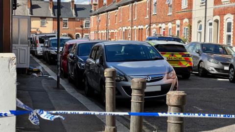 A residential street of terraced houses with a police cordon up around it. There's a police car stopped in the middle of the road.