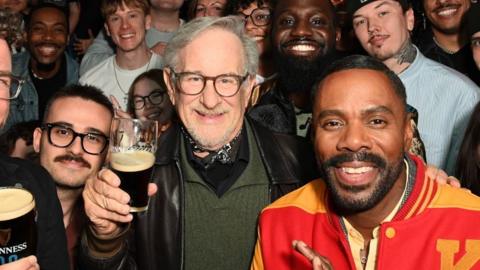 Steven Spielberg in a black leather jacket, green jumper and bandana around his neck, poses with fans inside a London pub while holding an half-full glass of Guinness. To his left is actor Colman Domingo in a red and yellow letterman jacket.