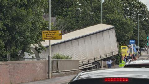 A lorry hangs off the edge of a road at a bridge as traffic builds. Someone in a hi-visibility vest is holding their hand up, directing traffic. There are trees lining the road as well as signs for businesses.