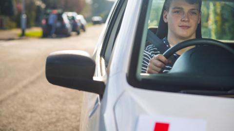 A male driver wearing a striped top sits in the driving seat of a car, with an L plate half visible. 