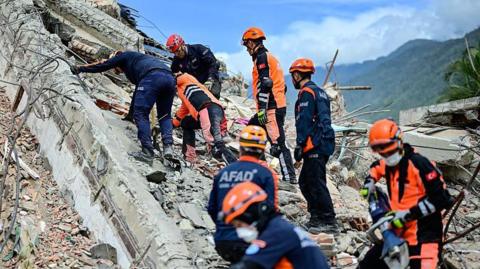 Turkish rescue teams in blue and orange work wear climb on top of building debris looking for survivors in La Guaira