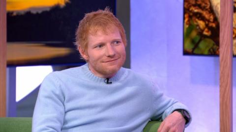 Ed Sheeran wearing a blue jumper while sitting on the One Show sofa. He's looking to the right and giving a small smile