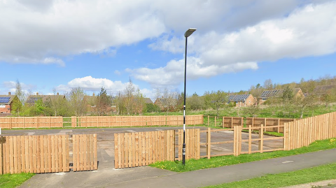 An empty patch of cement with light brown fencing around it. There are clouds and blue skies. There is a street light in the middle of the photo in front of the fencing. 