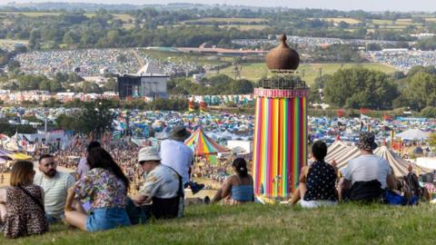 view overlooking Glastonbury festival with people sitting and enjoying the sunshine overlooking the site with the famous helter skelter