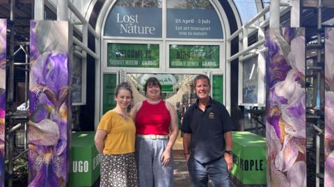 Left to right: Miffy Lane, Vikki Gray, and Chris Bichard. They are stood in front of Guernsey Museum. There are glass doors behind them and colourful displays around them.
