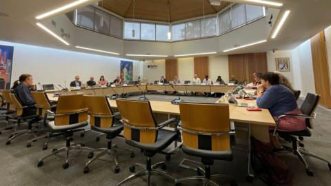 A council meeting in a white-walled room with wood panels and a large skylight. Councillors sit round a large square of desks.