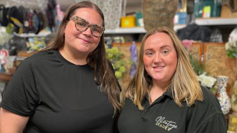 Two women wearing black branded T-shirts stand together in their store. The woman on the left is wearing speckled-rimmed glasses and has long brown hair. The woman on the right has shoulder-length fair hair.