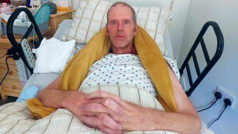 Tim Hull lying in an NHS hospital bed in his home. He is clasping his hands together and looking towards the camera. He has a yellow towel around his neck for support and is wearing a white t-shirt with blue spots.