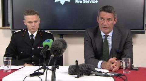 Oliver Lee and deputy Simon Barry sit at a desk in front of media microphones during a press conference.