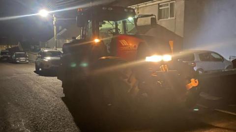 A tractor on a residential street stands with its lights on. 