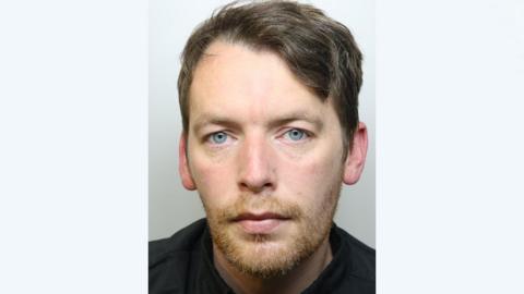 A police mugshot of Mark Manship wearing a black t-shirt and standing against a grey background. He has light blue eyes, short brown hair, and a ginger beard. 