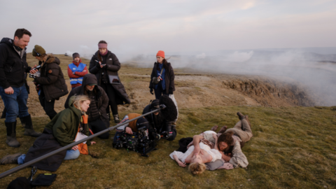 A scene of the 2026 film Wuthering Heights being shot in the Yorkshire Dales. To the left of the image are members of the crew with a boom microphone and camera. To the right Margot Robbie is lying on the ground in costume with Jacob Elordi to her side also in costume