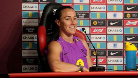 Lucy Bronze, wearing an England training vest, smiles as she speaks at a press conference