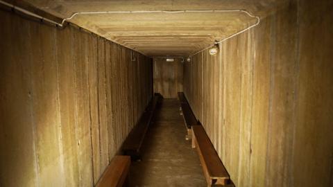 An underground tunnel. There are short, wooden benches either side and lights near the ceiling.