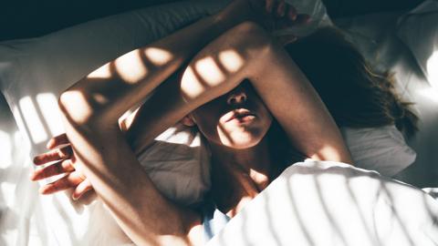 A woman lying in bed at home covering her eyes with her arms while sunlight streams in