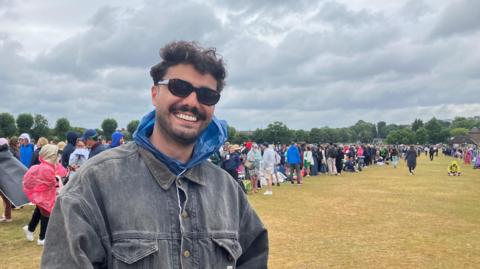A man in a grey denim jacket, big sunglasses and curly hair. 
