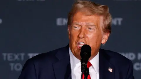 Donald Trump, wearing a blue suit and red tie, speaks into a microphone 