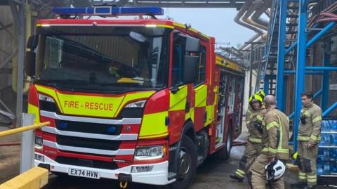 A red and yellow Essex Fire and Rescue truck in an industrial area. There are four firefighters in full gear standing next to it.