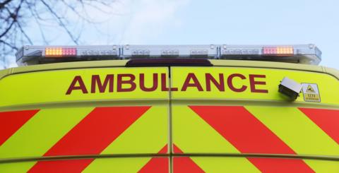 A close up of the back of an ambulance. It is yellow with orange stripes and 'AMBULANCE' is written in red text across the top of it.