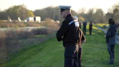 A side-on view of a police officer in black uniform, wearing a white and blue police flat cap, standing on the grassy bank of the River Nene looking down to the water. Other people are huddled behind him. There are trees in the background. It looks to be twilight.