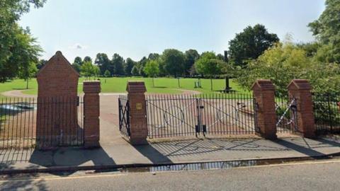 Black gates with brick pillars that open into a park which has a circular walkway and a stretch of green with various trees dotted about.