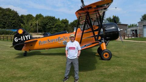 A man in a cap, white tshirt and grey trouser standing in front of an orange Tiger Moth plane