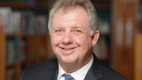 David Sidwick smiles at the camera. He has light grey hair, balding at the front, and wears a dark jacket and blue tie.