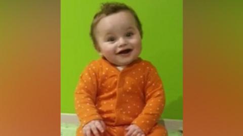 Undated image of the late baby Preston Davey. He has brown curly hair and brown eyes and is wearing an orange spotted baby grow. He is smiling.