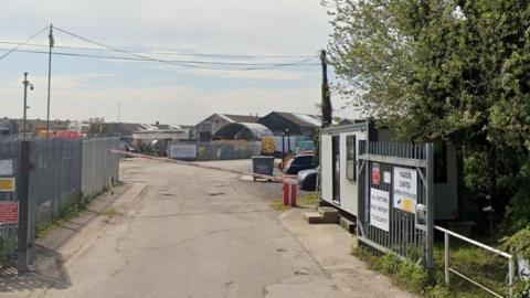 The entrance to an industrial estate, with metal gates to the fore and buildings in the background. 