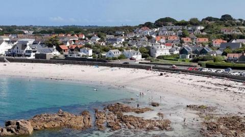 Guernsey with the sea in the corner and a beach. There are houses lined up nearby and a car park with cars parked next to the sand.