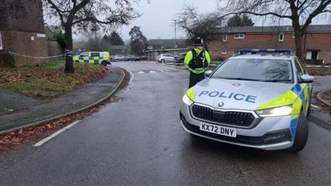 A parked police car with an officer standing on the driver's side next to some police tape in a housing estate. A police tape cordons off a house. 