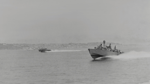 RON 30 boats speeding out of St Helier Harbour - black-and-white photograph showing two motor torpedo boats speeding across open water. Several crew members are visible as small figures on deck. A second boat follows at a distance toward the centre-left, also leaving a wake.