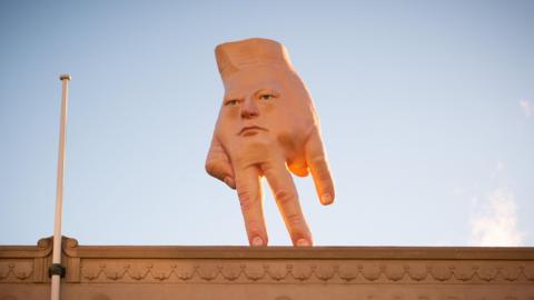 A statue of an upside down hand with its index and middle finger on the floor, the thumb is bent to look like a hand on hip. The ring finger is folded inwards to the pam and the pinky is outstretched. There is a human face on the back of the hand