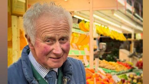 Robin Blair is standing in front of his fruit and vegetable stall and smiling at the camera. He has short white hair and rosy cheeks. He is wearing a blue body warmer and a green jacket underneath.
