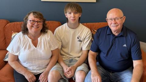Jacqui and Malcolm are sitting either side of Jack on a sofa. They are pensioner-age, with Jacqui wearing glasses and having shoulder-length, brown hair. Malcom is also wearing glasses and is bald with a moustache. Jack is a tall teenager with a fringe hairstyle and long hair at the back. The sofa is orange.