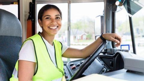 A woman bus driver sits behind the wheel and smiles at the camera. She is wearing a yellow high visibility jacket.